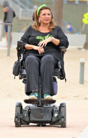 EXCLUSIVE: Dance Moms star Abby Lee Miller doesn’t flinch as a gymnast SOMERSAULTS over her during an outing to an LA beach. The reality TV star, 55, bravely lay back in her wheelchair as the muscular shirtless performer ran towards her then vaulted above her. Miller, who shot to fame as the star of hit show Dance Moms, has been using a wheelchair after undergoing surgery to remove a tumor from her spinal cord after reportedly being diagnosed with a form of cancer called Burkitt Lymphoma. Miller’s visit to the beach in Santa Monica, CA, saw her talking to a man and a woman dressed in workout gear before giving him a series of dance lessons. An onlooker said: “Abby was in great form and started giving the guy dance lessons. "She was teaching him to move and how and when to move his feet and it seemed he was struggling to get the hang of it. It was like she was back on the show Dance Moms and was shouting orders at him to ‘move to the side’ or ‘back right’. She appeared to be trying to teach him ballet just like she did to kids on the series. “It looked the guy had some gymnastic training and so afterwards they did a stunt where he ran at her and somersaulted directly above her.” Miller spent eight seasons on Dance Mom and previously taught Joelle Joanie “Jojo” Siwa. In 2017 she was sentenced to 366 days in prison for bankruptcy fraud. 31 Aug 2021 Pictured: Abbey Lee Miller. Photo credit: KAT/MEGA TheMegaAgency.com +1 888 505 6342 (Mega Agency TagID: MEGA782922_021.jpg) [Photo via Mega Agency]
