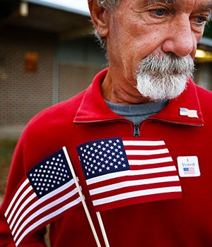 Man wearing an "I voted" sticker holding two American flags
