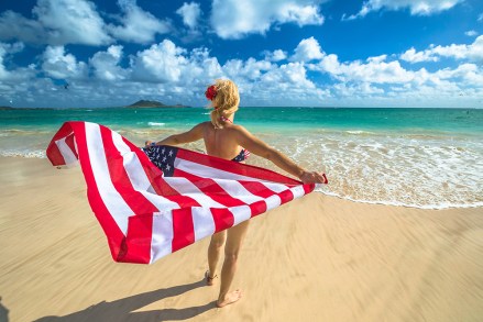 Back side Hawaiian woman holding a waving American flag in American flagged bikini. Tropical Lanikai Beach, east shore of Oahu in Hawaii, USA. Freedom and 4th July patriotic concept.; Shutterstock ID 1115229482; Comments: Art USe