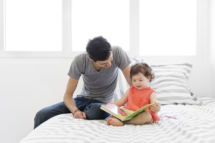 MODEL RELEASED Tattooed man wearing grey T-shirt and jeans and baby girl in red dress sitting on bed with stripy duvet, looking at book.
VARIOUS