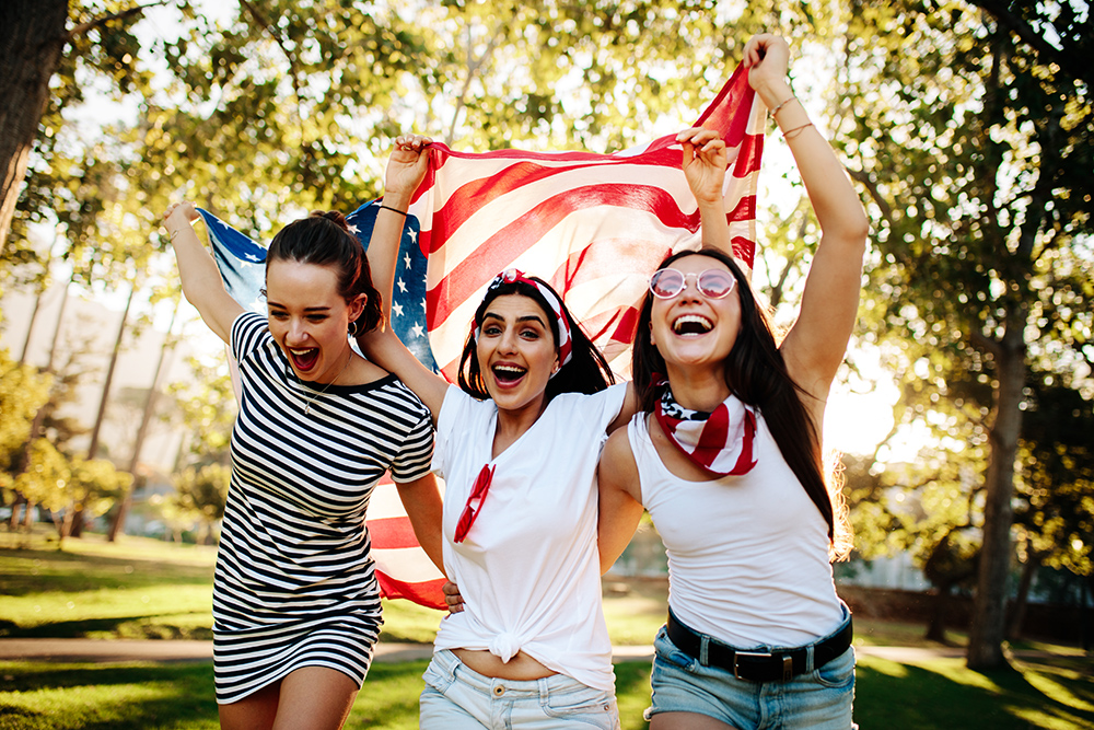 Cheerful female friends running around the park with American flag. Young and enthusiastic American girls enjoying 4th of july holiday at park.; Shutterstock ID 1377382046; Comments: art use