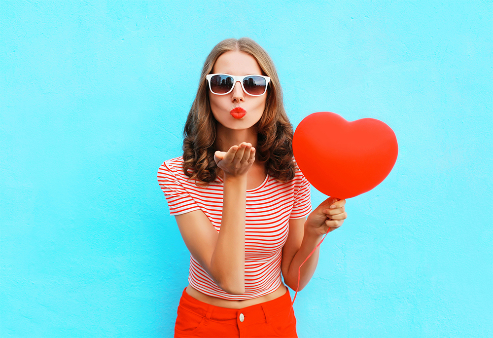 Portrait pretty woman sends air kiss with red balloon heart shape over blue background; Shutterstock ID 573140110; Comments: art use