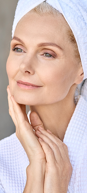 Happy beautiful middle aged woman wearing bathrobe and white towel with perfect complexion† touching face looking away in bathroom. Advertising of skin care spa wellness concept. Closeup portrait.