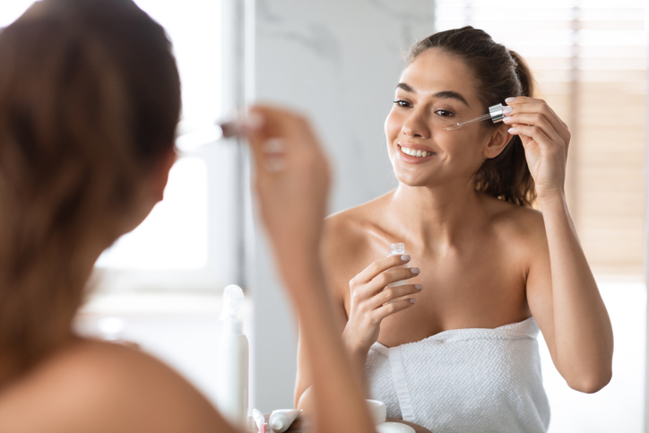 Facial Skincare. Young Woman Applying Face Serum Using Dropper Caring For Skin Standing Near Mirror In Modern Bathroom Indoors. Beauty Routine And Anti-Aging Skin Treatment Concept. Selective Focus