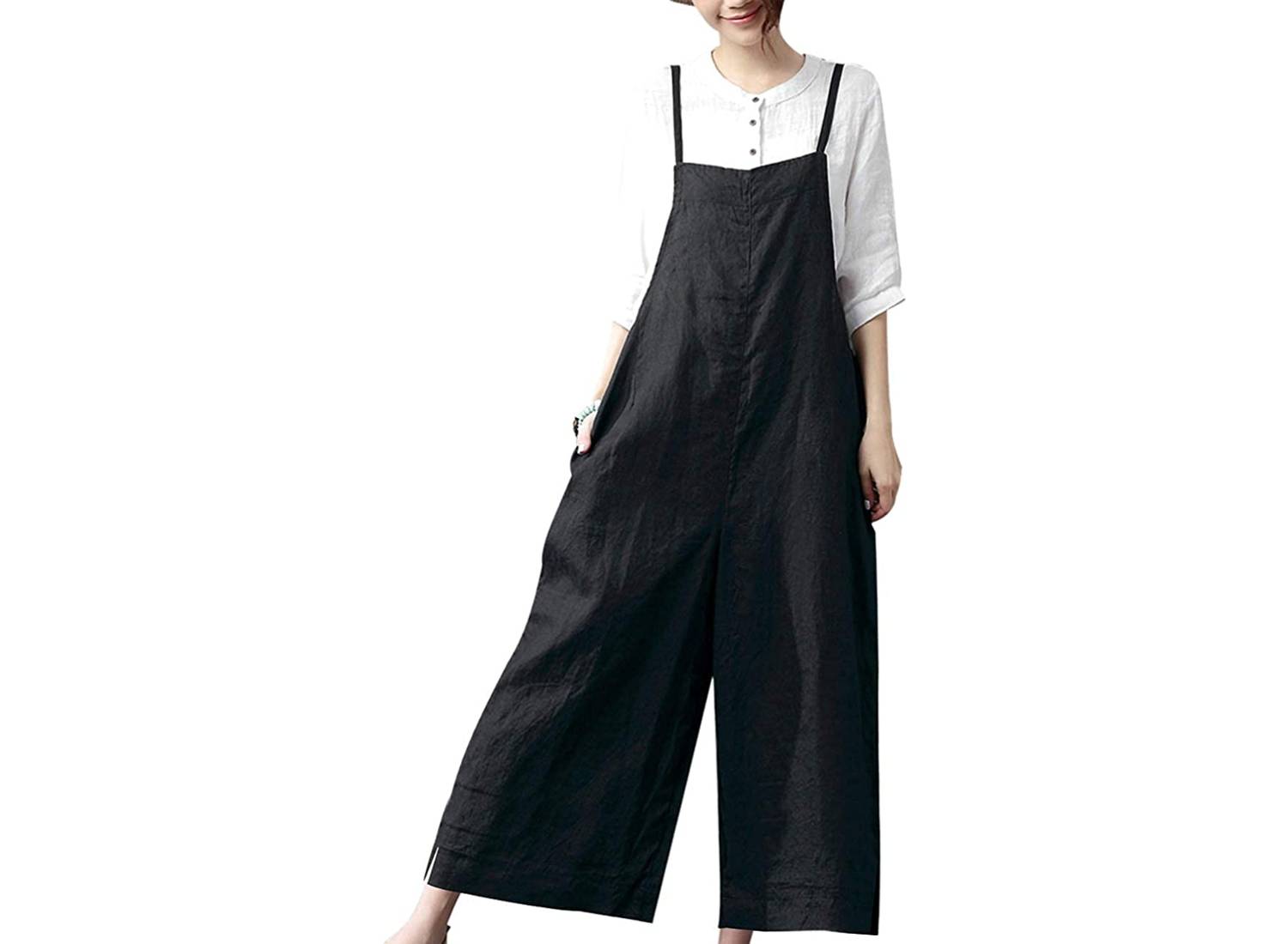 A woman wearing black, baggy wide-leg cotton ankle length overalls with a white long-sleeve shirt underneath 