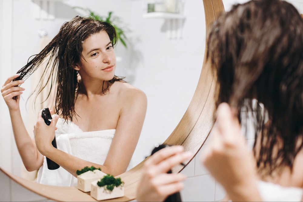 Young happy woman in white towel applying scalp cleanser on hair in bathroom, mirror reflection