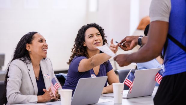 Two cheerful female volunteers help the unrecognizable young adult with his ballot for the election.