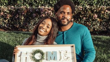 Smiling couple sitting on lawn holding a 'Home' sign, celebrating new home ownership.