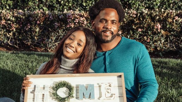 Smiling couple sitting on lawn holding a 'Home' sign, celebrating new home ownership.